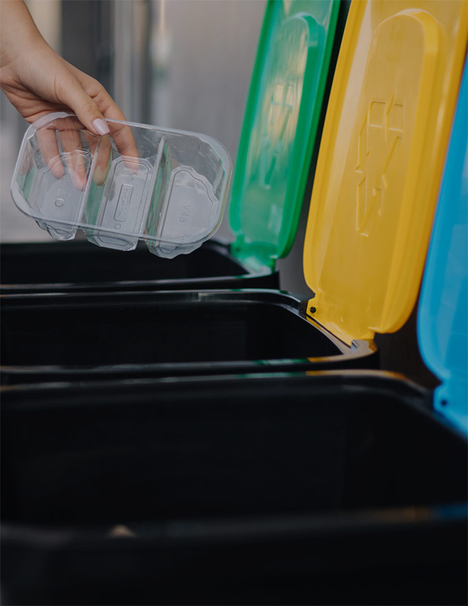 Photo de trois poubelles de tri une au couvercle vert, une au couvercle jaune, une au couvercle bleu. On voit une main déposer un emballage plastique dans la poubelle au couvercle jaune.