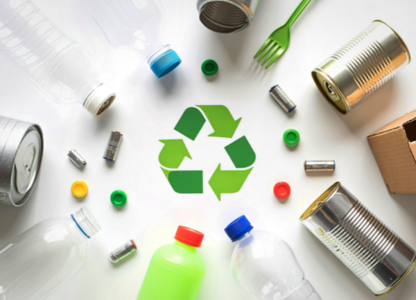 Photo montrant divers déchets recyclables disposés en cercle autour du symbole vert du recyclage : bouteilles en plastique transparent et coloré, bouchons, canettes métalliques, conserves, piles et un carton.