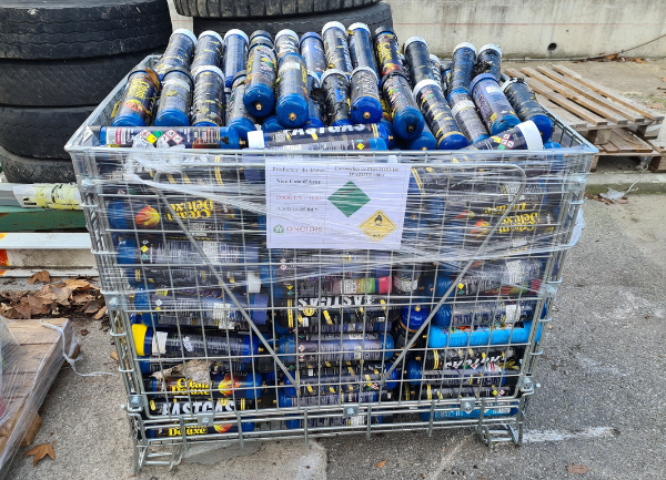 Grande caisse grillagée placée à l’extérieur sur un sol en béton, remplie de nombreuses cartouches cylindriques bleues (protoxyde d'azote). La caisse est entourée de film plastique transparent et contient plusieurs couches de ces cylindres empilés. À l’avant, une étiquette blanche est visible avec des pictogrammes de danger, dont un losange vert et un symbole de produit dangereux.