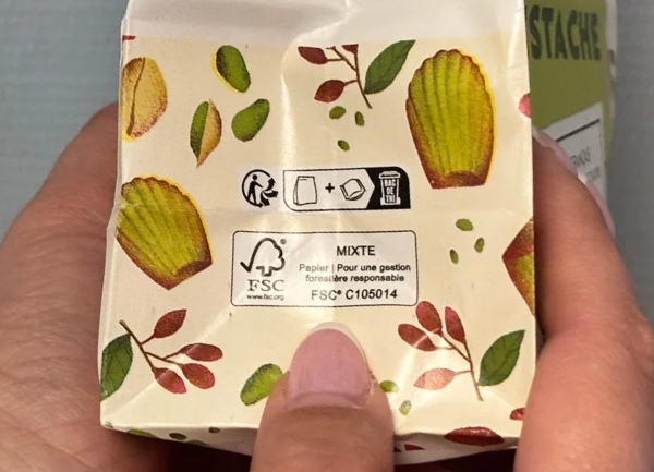 Emballage beige décoré de coquillages et de motifs végétaux colorés, tenu dans une main. Au dos, plusieurs pictogrammes de tri et un label FSC indiquant un emballage en matériaux mixtes.